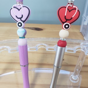 May include: Two pens with a heart-shaped charm with a stethoscope design. One pen is purple with glitter and the other is silver. Both pens have a white and red bead.