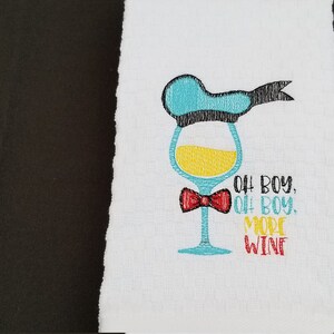 May include: A white kitchen towel with an embroidered design of a wine glass wearing a blue hat and a red bow tie. The text "Oh boy, Oh boy, More Wine" is embroidered below the glass.
