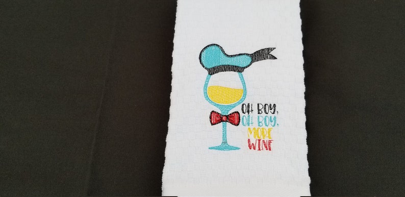 May include: A white kitchen towel with an embroidered design of a wine glass wearing a blue hat and a red bow tie. The text "Oh boy, Oh boy, More Wine" is embroidered below the glass.