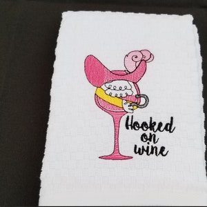 May include: A white kitchen towel with pink and yellow embroidery of a wine glass with a hook and the text "Hooked on wine".