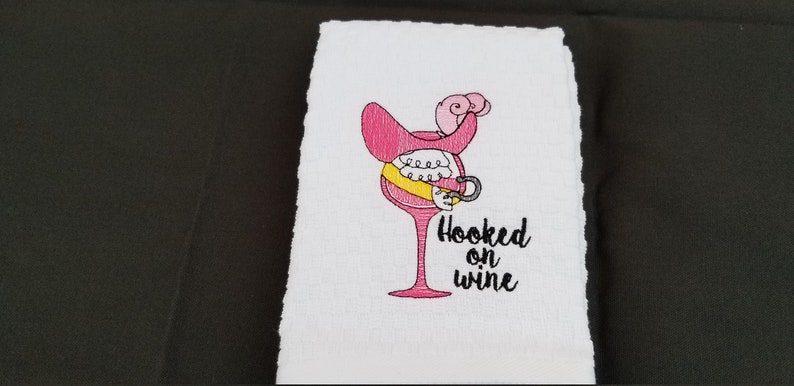 May include: A white kitchen towel with pink and yellow embroidery of a wine glass with a hook and the text "Hooked on wine".