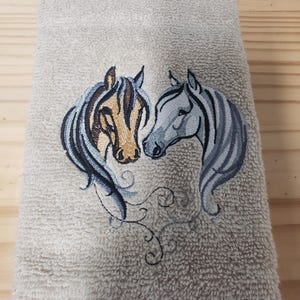 May include: Beige hand towel with an embroidered design of two horse heads facing each other. The horses are in shades of brown, gray, and white.