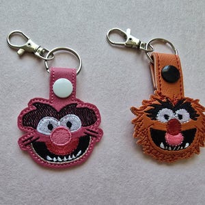 May include: Two embroidered keychains featuring cartoon character faces. One is pink with a white button, the other is brown with a black button. Both have silver-colored metal clasps and key rings. The keychains are likely made of felt or a similar material.