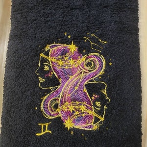 May include: Black towel with a gold and purple embroidered design of the Gemini zodiac sign. The sign is surrounded by stars and the symbol "II" is embroidered below.