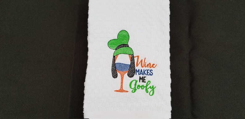May include: White kitchen towel with an embroidered design of a wine glass with a Goofy-like face and the text "Wine Makes Me Goofy".