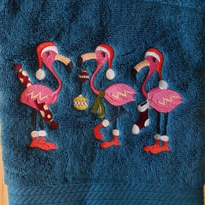 May include: Teal hand towel embroidered with three pink flamingos wearing Santa hats and holding Christmas ornaments and stockings. The flamingos have black beaks and legs, and are decorated with red, white, and green accents.