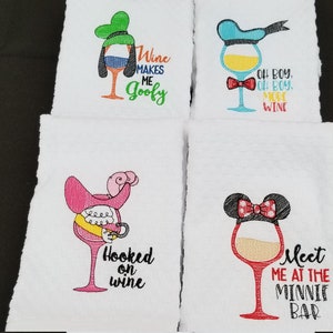 May include: Four white kitchen towels with embroidered wine glasses and Disney character designs. The designs include Goofy, Donald Duck, Minnie Mouse, and a pink flamingo. The towels have text that reads "Wine Makes Me Goofy", "Oh Boy, Oh Boy, Wine", "Hooked on Wine", and "Meet Me at the Minnie Bar".