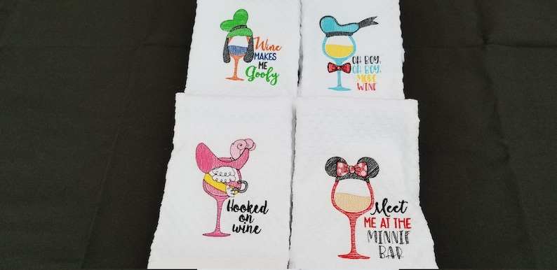 May include: Four white kitchen towels with embroidered wine glasses and Disney character designs. The designs include Goofy, Donald Duck, Minnie Mouse, and a pink flamingo. The towels have text that reads "Wine Makes Me Goofy", "Oh Boy, Oh Boy, Wine", "Hooked on Wine", and "Meet Me at the Minnie Bar".