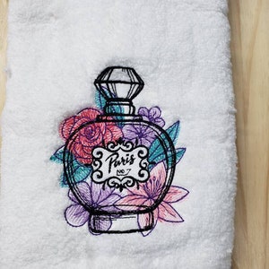 May include: Embroidered white hand towel with a black and white outline of a perfume bottle with a diamond-shaped stopper. The bottle is surrounded by pink, purple, and teal flowers. The bottle has a label that says "Paris No. 7".