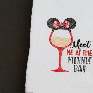 May include: White kitchen towel with red embroidery of a wine glass with a Minnie Mouse ear and bow on top. The text "Meet me at the Minnie Bar" is embroidered below the glass.