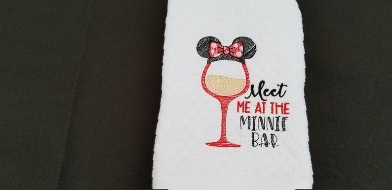 May include: White kitchen towel with red embroidery of a wine glass with a Minnie Mouse ear and bow on top. The text "Meet me at the Minnie Bar" is embroidered below the glass.