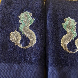 May include: Two dark blue bath towels with embroidered mermaid designs. The mermaids are silver with green hair and tails.