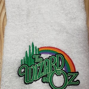 May include: White towel with an embroidered design featuring the words "The Wizard of Oz" in green and black, a green Emerald City, and a rainbow. The towel is folded and has a textured surface.