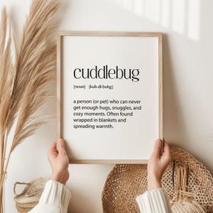 May include: A framed print with the word "cuddlebug" in a black, modern font. The definition below describes a person who loves hugs and cozy moments. The frame is light-colored, and the background is white.