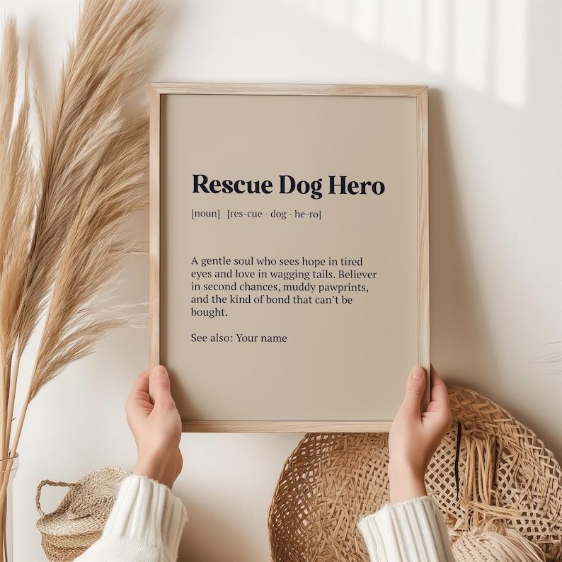 Op de afbeelding: Een ingelijste print met de tekst "Rescue Dog Hero" in een donkerblauwe lettertype. De print bevat een definitie en een beschrijving van een reddingshond. De achtergrond is lichtbruin en de lijst is licht hout.