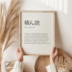May include: A framed print with a black and white design. The print features the Japanese word "積ん読" (tsundoku) and its definition. The definition explains that tsundoku is the habit of buying books and letting them accumulate, unread, on shelves. It celebrates the love of books and the anticipation of future reading.