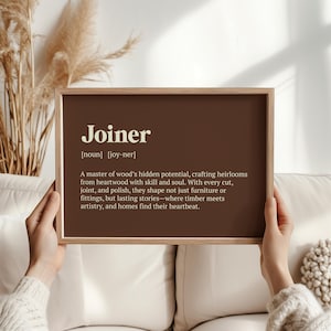 May include: A framed print with the definition of "Joiner" in white text on a brown background. The definition describes a joiner's skill in crafting heirlooms from wood. The frame is a light brown color.