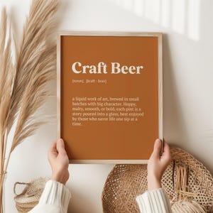 May include: A framed print with the words "Craft Beer" in white text on a burnt orange background. The definition of craft beer is below the title. The print is held by a person.