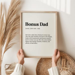 May include: A framed print with a white background and black text that reads "Bonus Dad [noun] [boh-nuhs - dad] Not just a dad, but a bonus in every way. The one who teaches you how to fix things, helps with homework, and knows exactly how to cheer you up. He's the extra father figure who brings warmth, wisdom, and plenty of dad jokes."