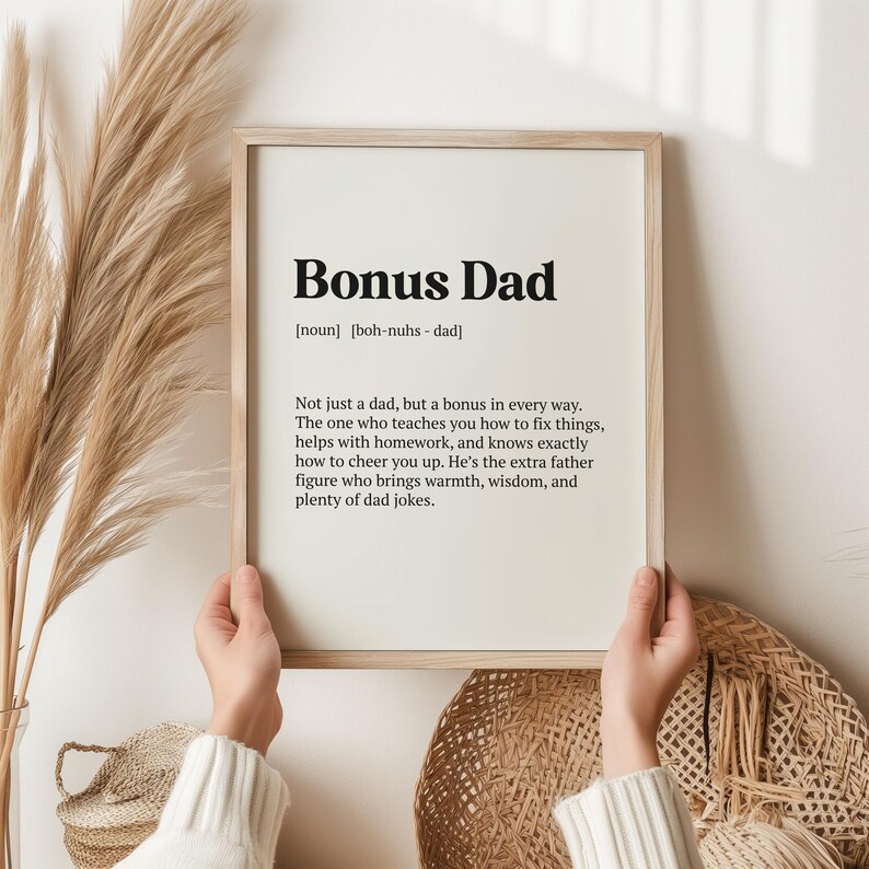 May include: A framed print with a white background and black text that reads "Bonus Dad [noun] [boh-nuhs - dad] Not just a dad, but a bonus in every way. The one who teaches you how to fix things, helps with homework, and knows exactly how to cheer you up. He's the extra father figure who brings warmth, wisdom, and plenty of dad jokes."