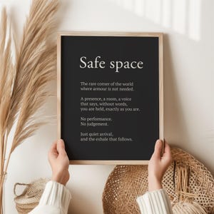 May include: A framed black print with the words "Safe space" in white. The text below describes a place of comfort and acceptance. The print is held by a person in front of a neutral background with decorative elements.