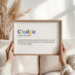May include: A framed print with the word "Cludgie" in blue and yellow, a Scottish term for toilet. The definition is printed below in black text. The frame is a light brown color.
