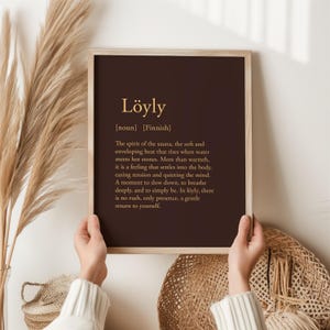 May include: A framed print with the word "L&ouml;yly" in gold lettering on a dark brown background. The text below describes the Finnish word's meaning, relating to the spirit of the sauna. The frame is light brown.