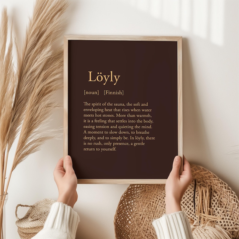 May include: A framed print with the word "L&ouml;yly" in gold lettering on a dark brown background. The text below describes the Finnish word's meaning, relating to the spirit of the sauna. The frame is light brown.