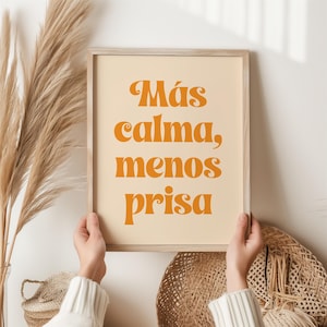 Op de afbeelding: Een ingelijste print met de tekst "Más calma, menos prisa" in oranje script op een crèmekleurige achtergrond. De lijst is van licht hout. De print wordt vastgehouden door een persoon die een witte trui draagt. Decoratief pampasgras en een rieten mand staan op de achtergrond.
