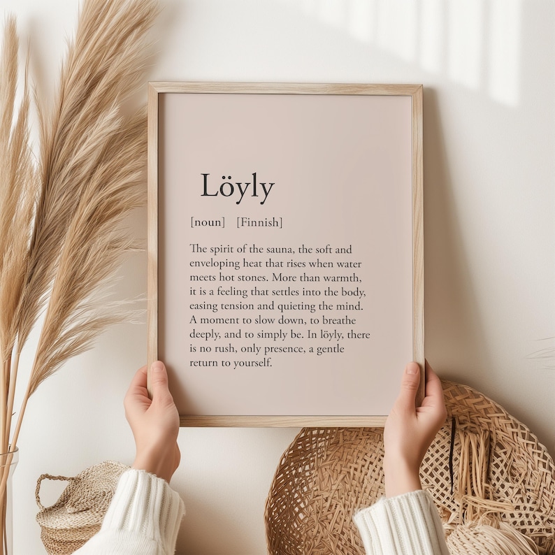 May include: A framed print with the word "L&ouml;yly" in a serif font, with a definition below. The print has a light wood frame and a neutral background. The text describes the spirit of the sauna. The print is held by a person.