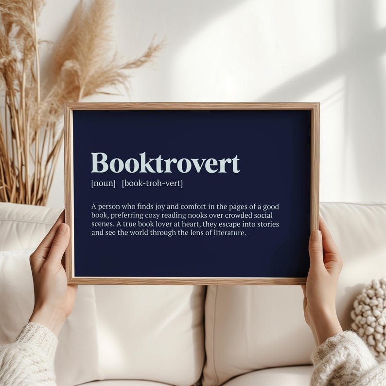 May include: A navy blue printable wall art print with the definition of the word "Booktrovert" in white text. The definition reads: "A person who finds joy and comfort in the pages of a good book, preferring cozy reading nooks over crowded social scenes. A true book lover at heart, they escape into stories and see the world through the lens of literature."