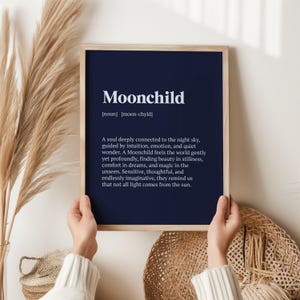 May include: A framed print featuring the word "Moonchild" in white against a dark blue backdrop. The definition of Moonchild is also printed below the word. The frame is a light wood colour.