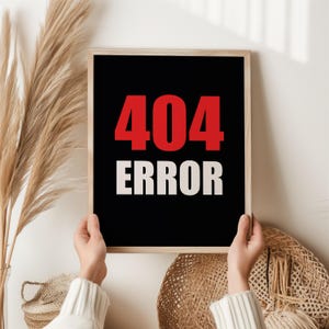 Könnte beinhalten: Ein gerahmter Kunstdruck mit schwarzem Hintergrund und den Worten "404 ERROR" in Rot und Weiß. Der Druck wird von einer Person in einem weißen Pullover gehalten. Das Kunstwerk wird in einem hellfarbigen Holzrahmen präsentiert.