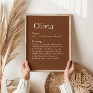 May include: A framed print with the name "Olivia" in a serif font against a brown background. The print includes the origin and meaning of the name, with text in a white font. The frame is a light wood color.