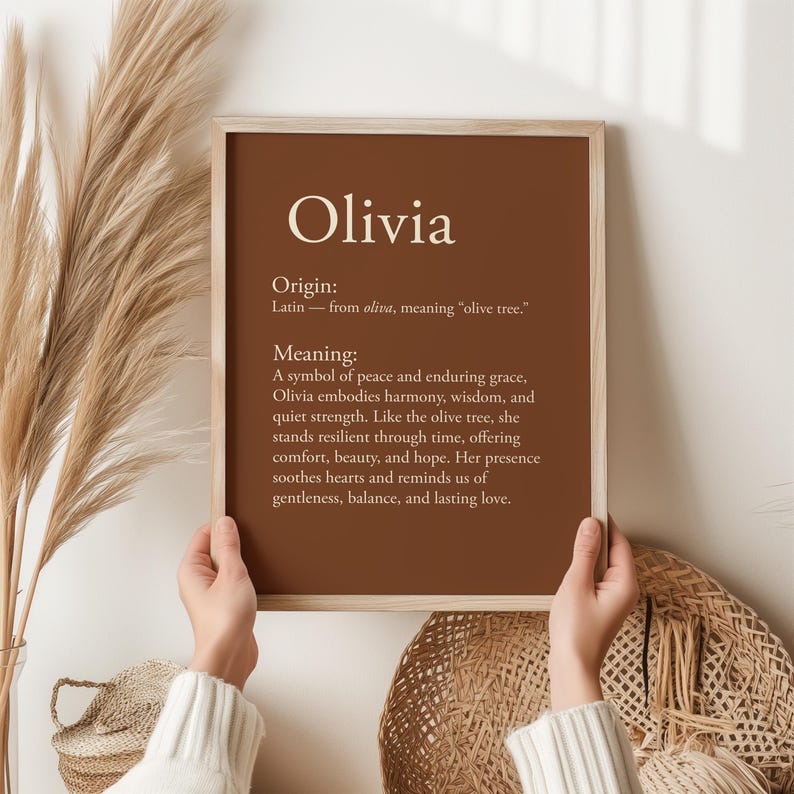 May include: A framed print with the name "Olivia" in a serif font against a brown background. The print includes the origin and meaning of the name, with text in a white font. The frame is a light wood color.