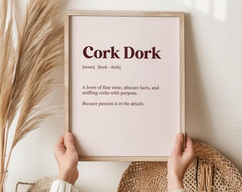 Op de afbeelding: Een ingelijste print met de tekst "Cork Dork" in bordeauxrood. De definitie beschrijft een liefhebber van fijne wijn, obscure feiten en het ruiken aan kurken. De print wordt door een persoon vastgehouden.