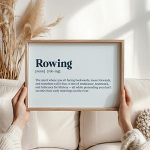 May include: A framed print featuring the definition of "Rowing". The text is in a dark blue font on a white background, set within a light brown wooden frame. The definition humorously describes the sport's challenges and teamwork.