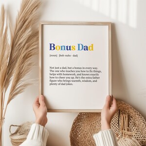 May include: A framed print with a white background and black text that reads "Bonus Dad" with a definition of the word. The text is in blue, yellow, and orange.