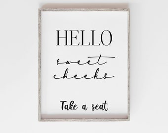 May include: A framed print with the text "HELLO sweet cheeks Take a seat" in black lettering on a white background.