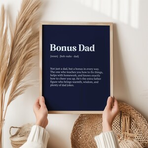 May include: A framed print with a navy blue background and white text that reads "Bonus Dad [noun] [boh-nuhs - dad] Not just a dad, but a bonus in every way. The one who teaches you how to fix things, helps with homework, and knows exactly how to cheer you up. He's the extra father figure who brings warmth, wisdom, and plenty of dad jokes."