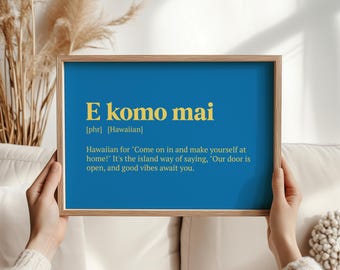 Op de afbeelding: Een ingelijste print met de woorden "E komo mai" in gouden letters op een blauwe achtergrond. De tekst hieronder legt de Hawaiiaanse zin uit, die "Kom binnen en maak het je gemakkelijk!" betekent.