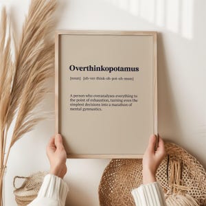 May include: A beige framed print with the word "Overthinkopotamus" defined as "A person who overanalyses everything to the point of exhaustion, turning even the simplest decisions into a marathon of mental gymnastics."