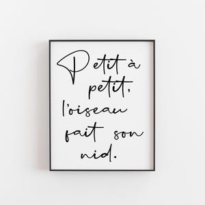 May include: A framed print with the French phrase "Petit à petit, l'oiseau fait son nid" in black script. The artwork is displayed in a black frame against a white background, suitable for home decor.