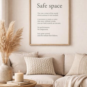May include: A framed print with the words "Safe space" in a minimalist font. The print is displayed above a beige sofa with decorative pillows and a wooden coffee table. A vase with dried pampas grass and a lit candle add to the cozy atmosphere.