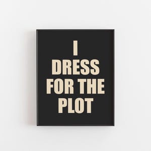 May include: A framed black print with the text "I DRESS FOR THE PLOT" in a beige, bold font. The print is rectangular and has a simple, modern design, suitable for home decor.