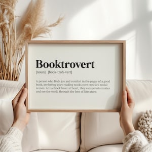 May include: A printable definition of the word "Booktrovert" in a white frame with a wood-toned border. The definition reads: "A person who finds joy and comfort in the pages of a good book, preferring cozy reading nooks over crowded social scenes. A true book lover at heart, they escape into stories and see the world through the lens of literature."