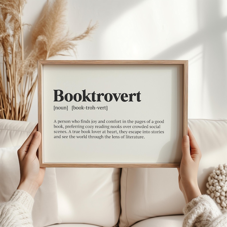 May include: A printable definition of the word "Booktrovert" in a white frame with a wood-toned border. The definition reads: "A person who finds joy and comfort in the pages of a good book, preferring cozy reading nooks over crowded social scenes. A true book lover at heart, they escape into stories and see the world through the lens of literature."