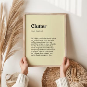 May include: A framed wall art print with a light yellow background featuring the word Clutter in bold black text, followed by its phonetic pronunciation and a dictionary-style definition describing the accumulation of objects in a home.