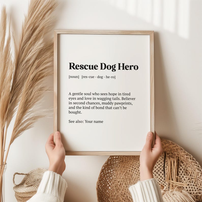 Op de afbeelding: Een ingelijste print met de tekst "Rescue Dog Hero" in zwarte letters. De definitie bevat een beschrijving van reddingshonden. De print wordt door handen vastgehouden, met een neutrale achtergrond.