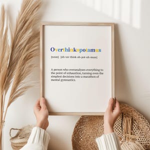 May include: A framed print with a light brown wood frame and a white background. The print features the word "Overthinkopotamus" in blue and yellow letters with a definition below. The definition reads: "[noun] [oh-ver-think-oh-pot-uh-muss] A person who overanalyses everything to the point of exhaustion, turning even the simplest decisions into a marathon of mental gymnastics."
